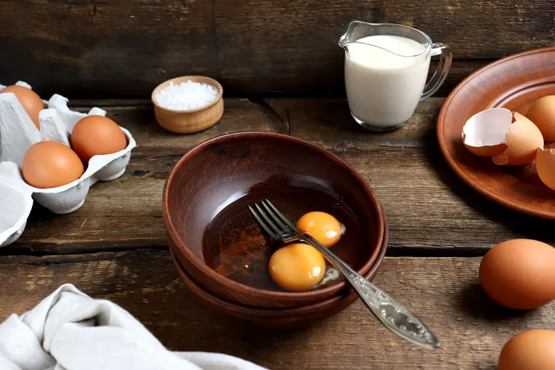 Brown eggs, a cracked egg in a bowl and a jug of milk on a rustic wooden table