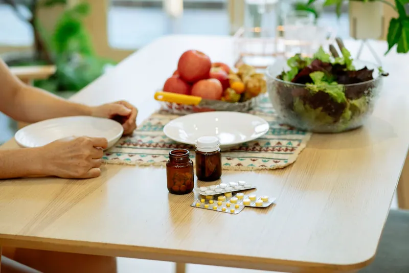 Supplement bottles and blister pack on a table next to a bowl of salad greens and fresh apples